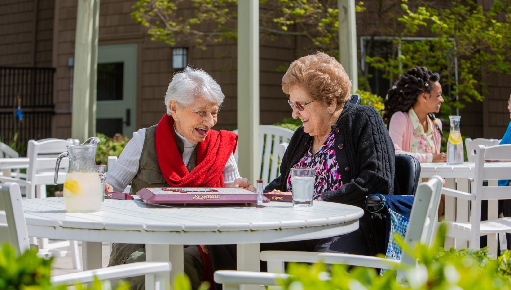Two elderly women sit at a round outdoor table, laughing while playing Scrabble. There are glasses of water and a pitcher with lemon slices on the table. Other people are sitting and talking in the background on a sunny day.