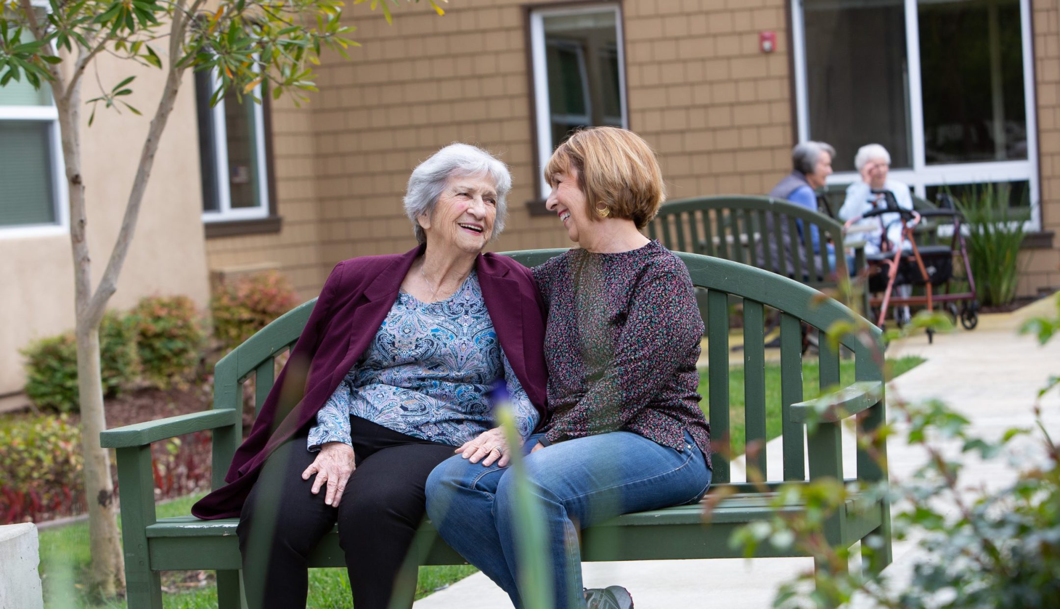 Two women sit closely together on a green bench outside, smiling and laughing. In the background, two other elderly women are sitting and talking near a building. The scene appears warm and friendly.