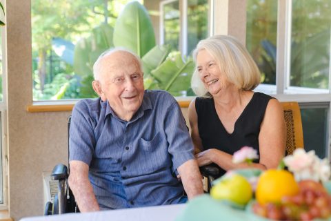 An elderly man in a wheelchair sits beside a smiling woman with gray hair at Eskaton Gold River senior living. Indoors, near a window and green plants, they enjoy each other’s company. A table with fruit is in the foreground.
