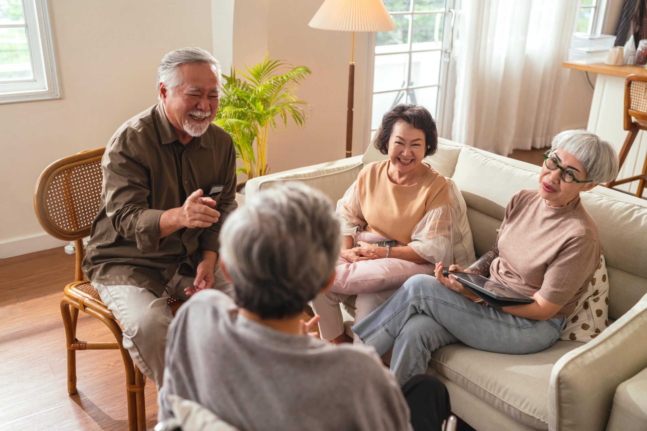 Four older adults sit together in a bright living room, smiling and talking. Like many dementia support groups, they share stories as one man points while the others laugh, creating a warm atmosphere with sunlight streaming through large windows.