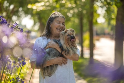 An older woman with long gray hair smiles while holding a small, fluffy dog outdoors. Surrounded by purple flowers and trees in warm sunlight, she enjoys exploring different living options for her golden years.