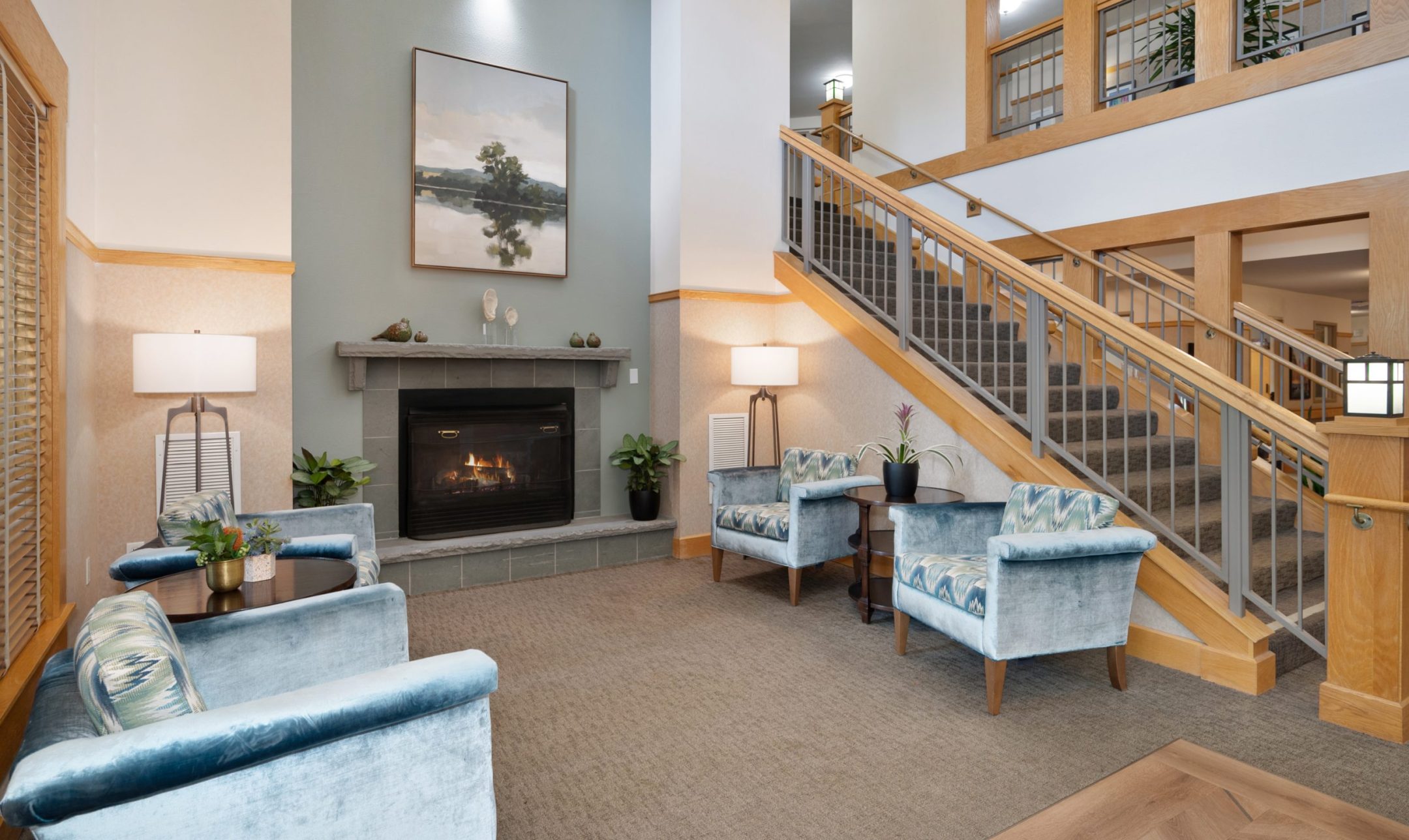 A cozy lounge area with blue armchairs, a fireplace, a large landscape painting above the mantel, side tables with lamps and plants, and a wooden staircase with a metal railing leading to an upper floor.