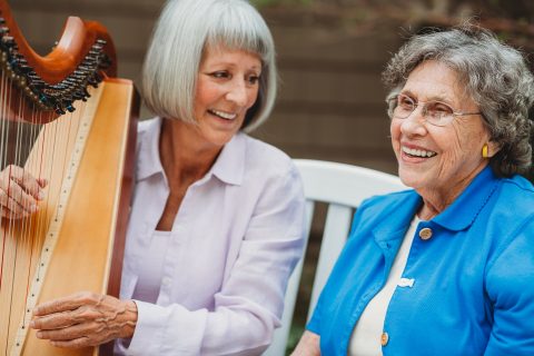 Two older women sit together smiling outdoors; one, with short gray hair, plays a harp while the other, wearing glasses and a blue jacket, listens joyfully.
