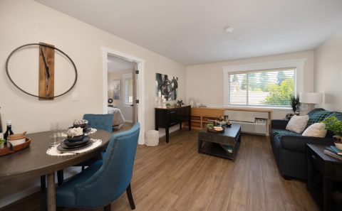 A modern living and dining area with wood flooring, teal chairs, a dark sofa, a coffee table, and large windows letting in natural light. A bedroom is visible through a sliding door on the left.