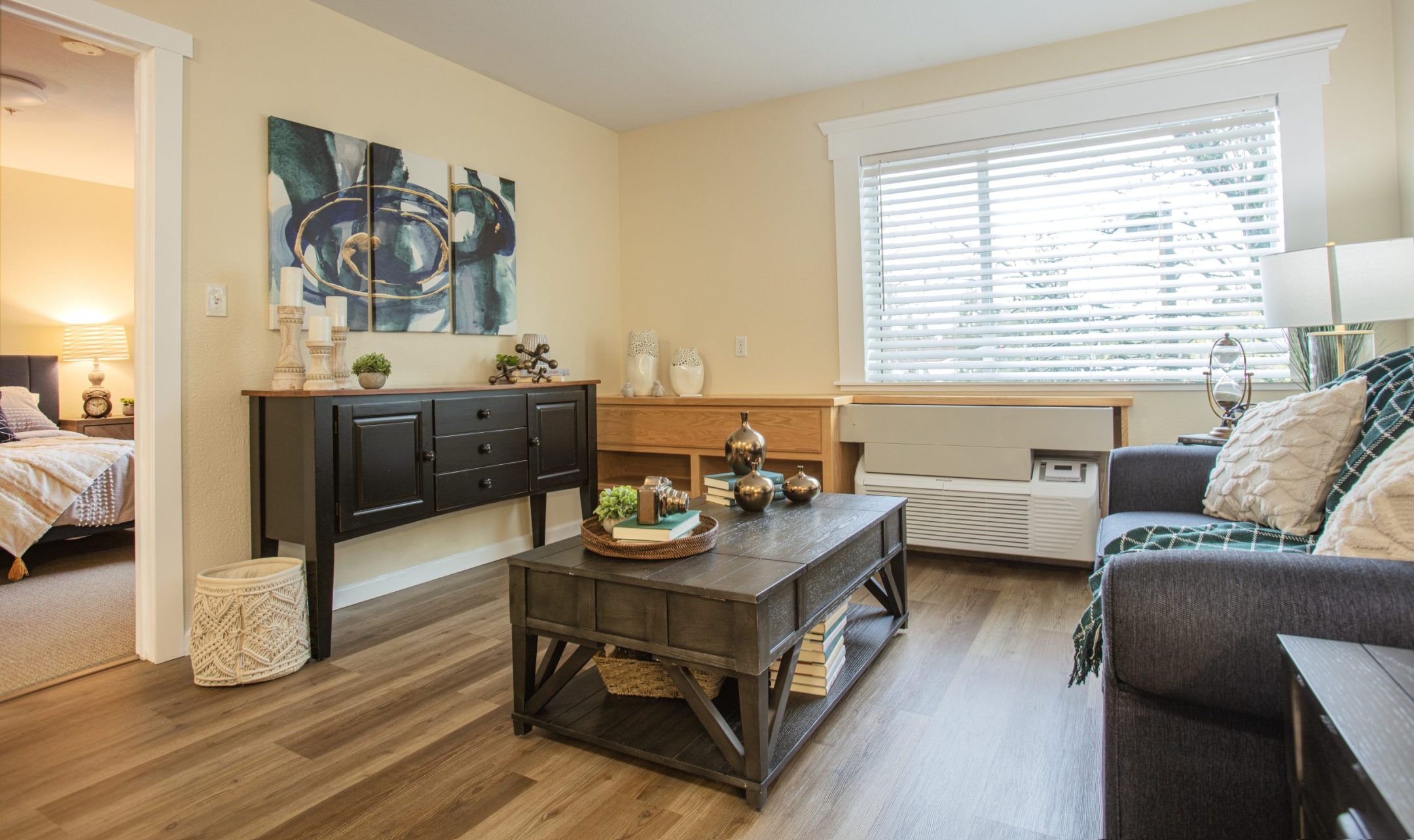 A cozy living room reflecting the Eskaton Gold River lifestyle features a dark wooden coffee table, a sofa, a black sideboard, decor items, and a large window with blinds. The adjacent bedroom is partially visible through an open doorway.