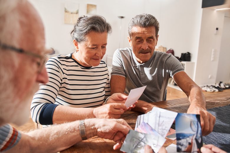 Three older adults sit at a table indoors in Eskaton Gold River memory care, sharing photographs together. They appear engaged and nostalgic, enjoying each other's company in a bright, cozy room.
