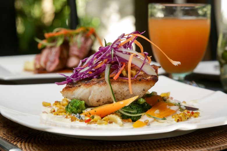 A gourmet dish from Eskaton Gold River dining features a seared fish fillet topped with shredded purple cabbage and carrots, served on a bed of vegetables, with a glass of orange beverage in the background.