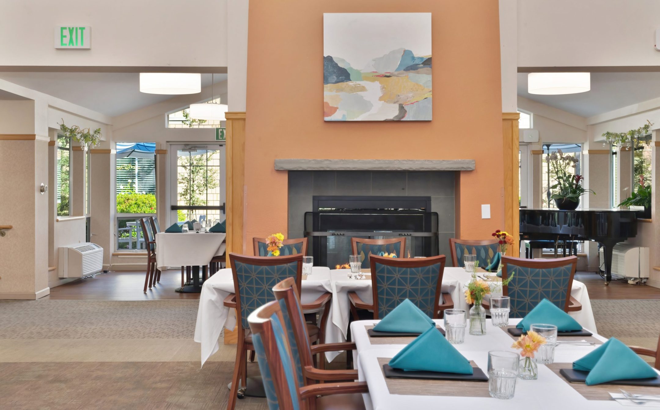 The Eskaton Gold River dining area features tables set for a meal with blue napkins and flower centerpieces. A fireplace with abstract art above it and a grand piano near large windows complete the inviting atmosphere.