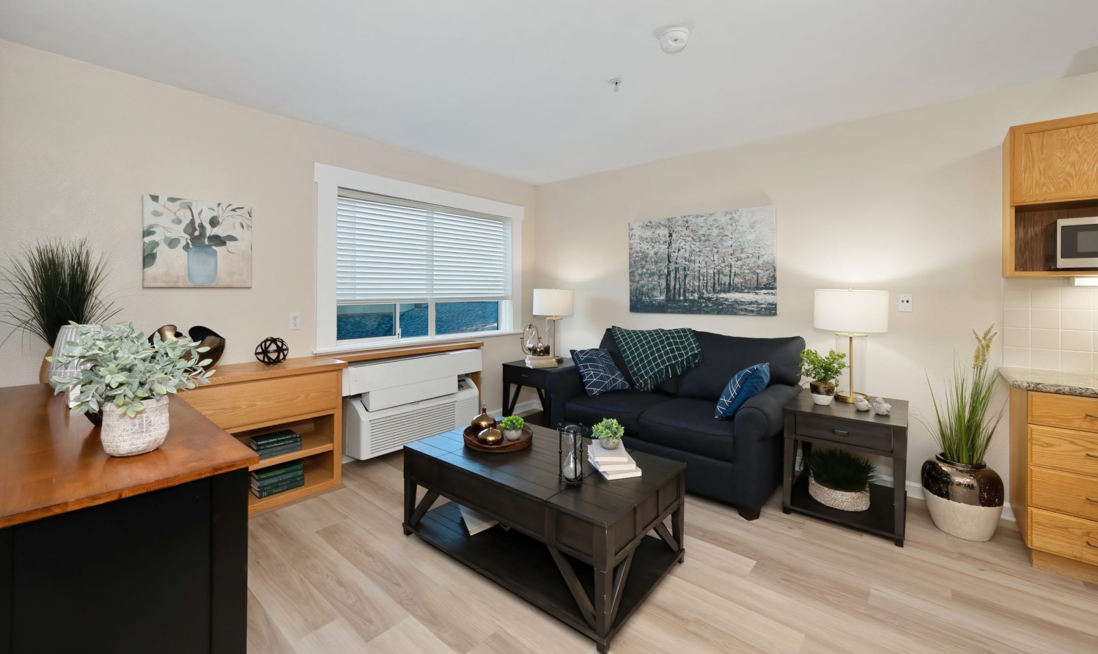 A cozy living room at Eskaton Gold River with a dark blue sofa, green cushions, black coffee table, indoor plants, art, and large window with blinds letting in natural light. Enjoy modern decor and inviting living options with light wood flooring.