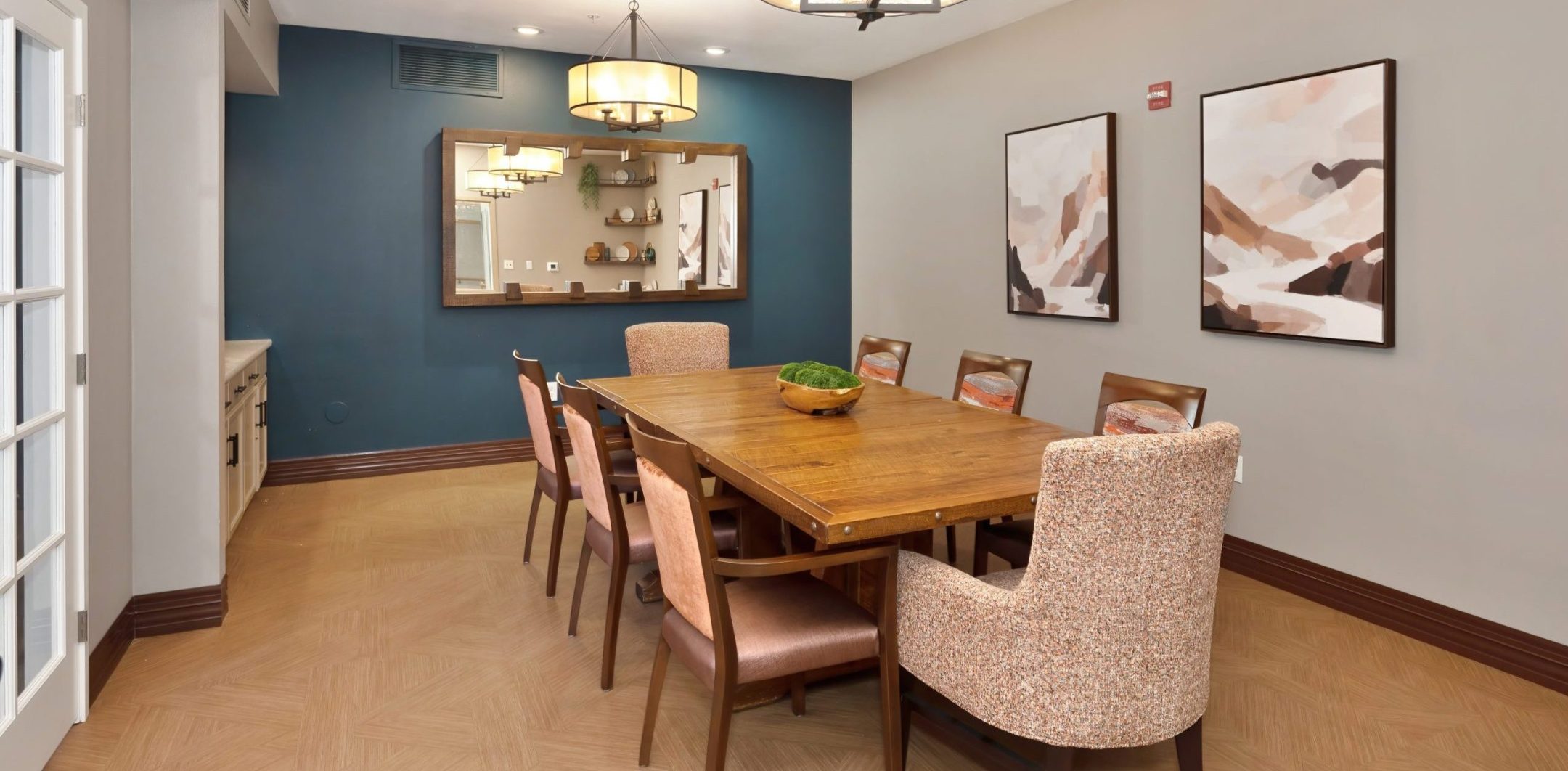 A modern Eskaton Gold River dining room features a wooden table, eight chairs, abstract paintings on a gray wall, a large mirror, and two round ceiling lights. Light wood floors complement the dark blue accent wall for an inviting ambiance.