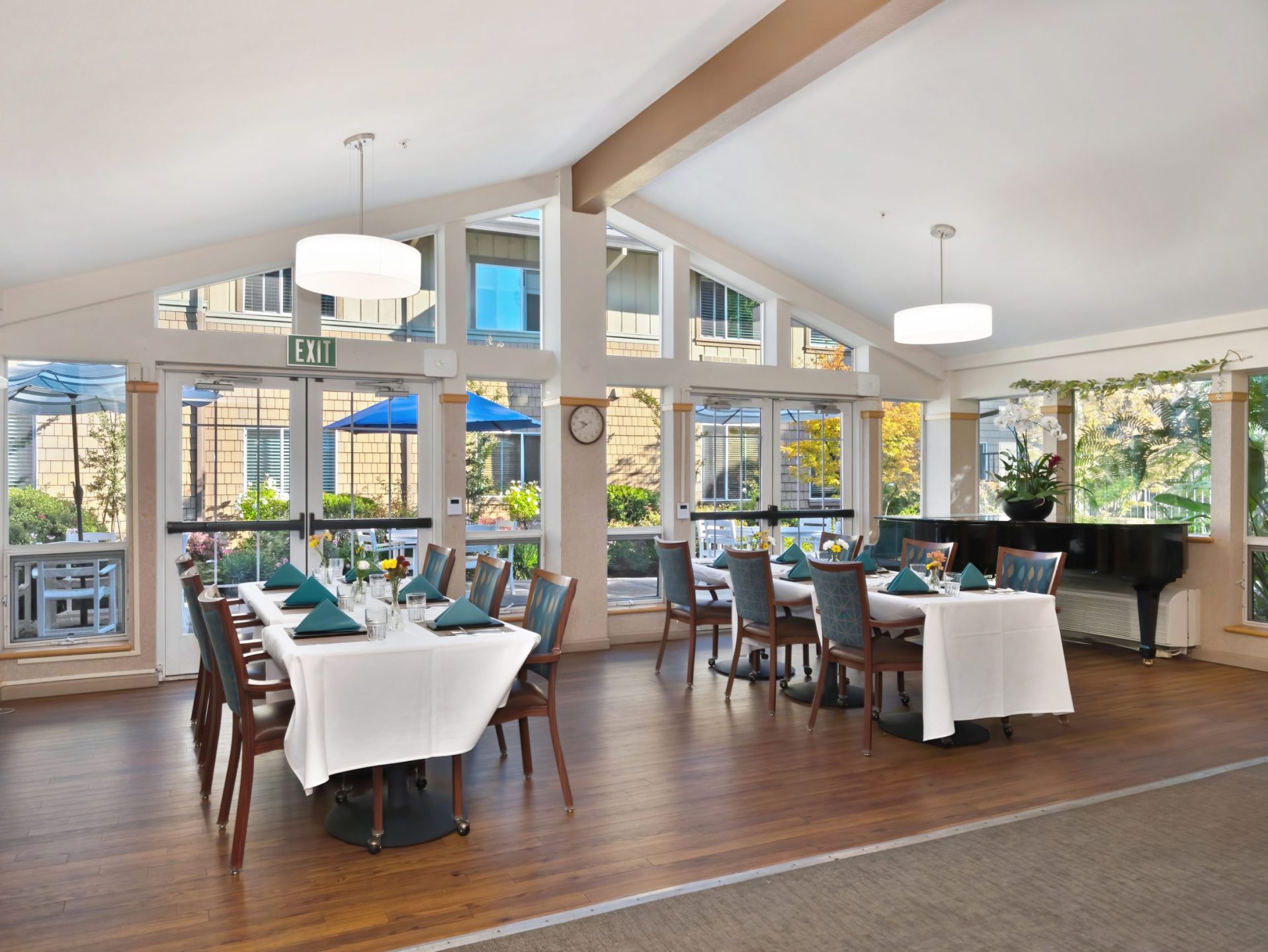 A bright, modern dining room at Eskaton Gold River assisted living features large windows, set tables with green napkins, and wooden chairs. Outside, blue patio umbrellas add color. Indoors, a black piano and plants bask in sunlight.