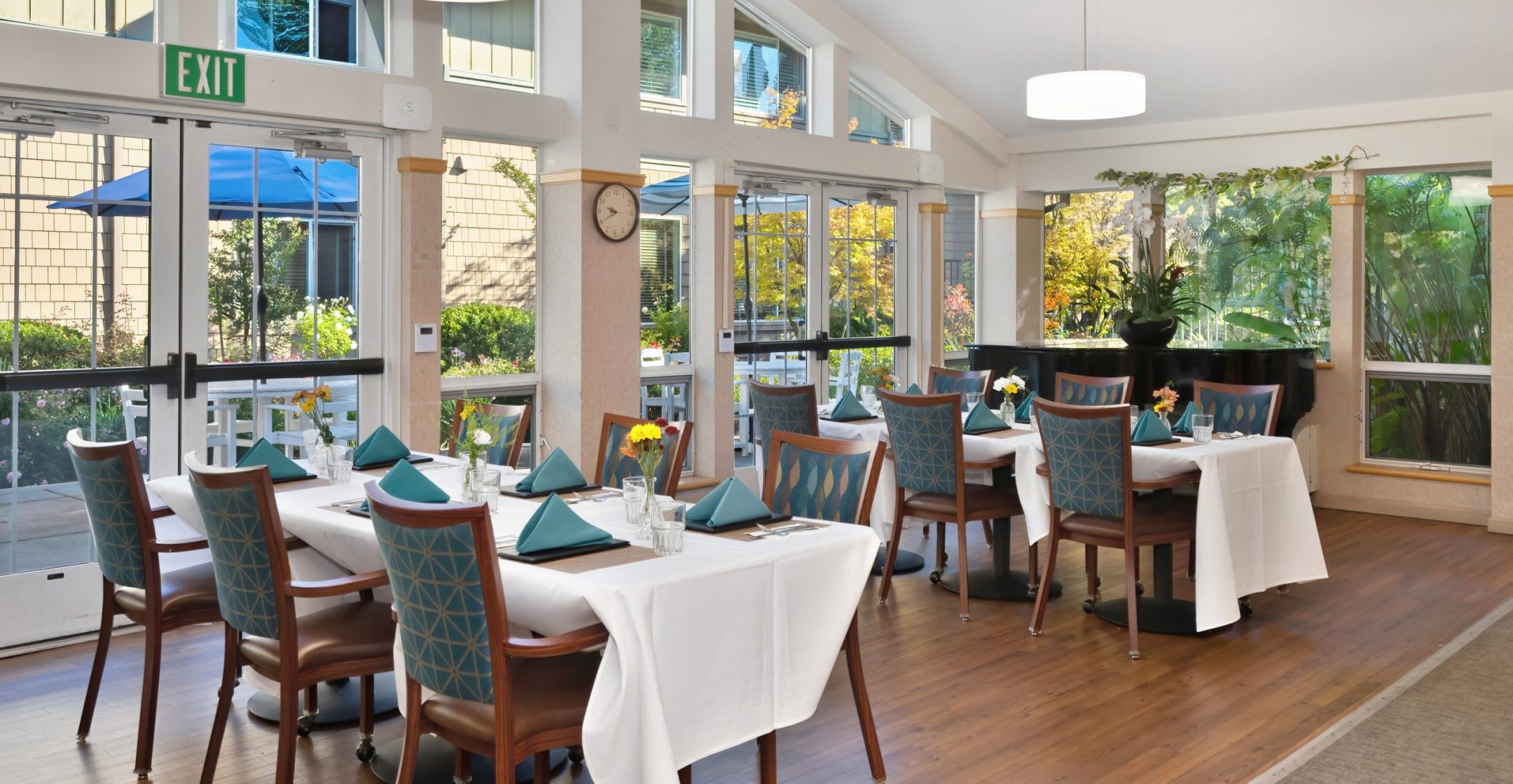 A bright Eskaton Gold River dining area with large windows, set tables with teal napkins, and wooden chairs. Flowers grace each table, modern hanging lights illuminate the space, and there's a sunny outdoor patio with blue umbrellas.