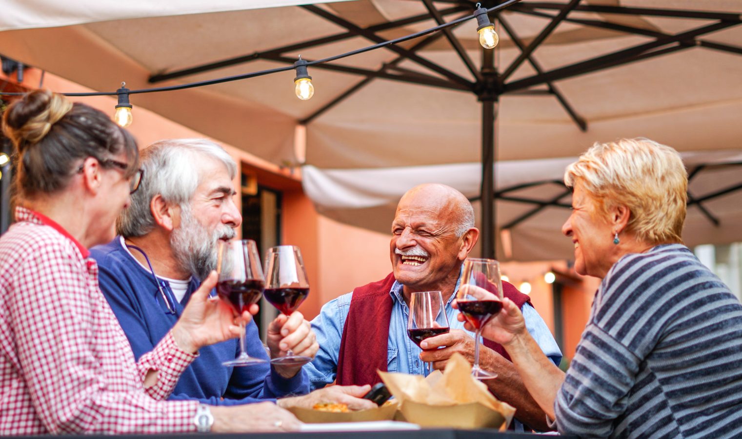 Four older adults sit at an outdoor table under umbrellas, smiling and clinking glasses of red wine, enjoying each others company. Warm lighting and casual attire create a relaxed, joyful atmosphere.
