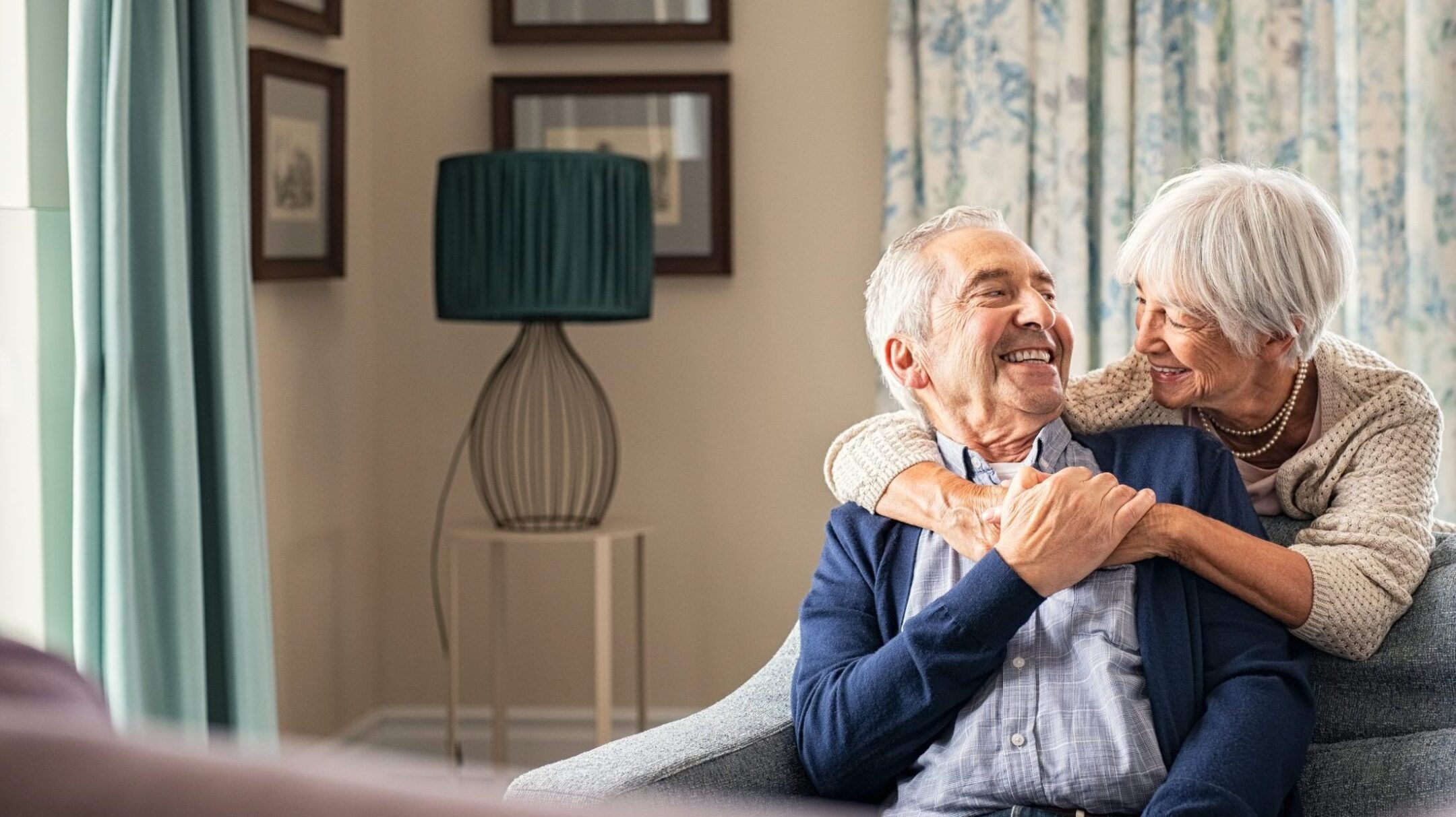 An elderly couple sits on a couch at home, smiling and embracing warmly, with the woman hugging the man from behind. The room is softly lit with patterned curtains and framed pictures on the wall.