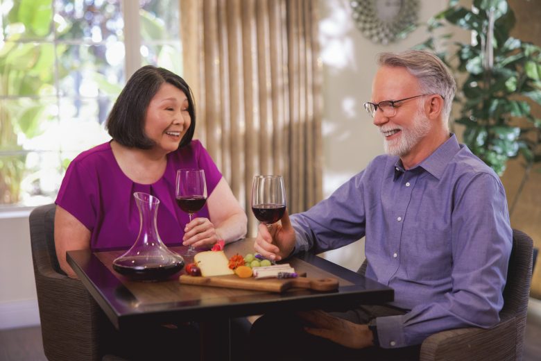 An older woman and man sit at a small table, smiling and holding glasses of red wine. A decanter, cheese, and fruit are on the table. They appear to be enjoying a pleasant conversation in a bright, cozy room.