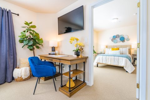A bright, tidy bedroom and workspace with a wooden desk, blue chair, potted plant, wall-mounted TV, decorative plants, and a bed with colorful pillows—showcasing flexible Living Options in the adjoining room.