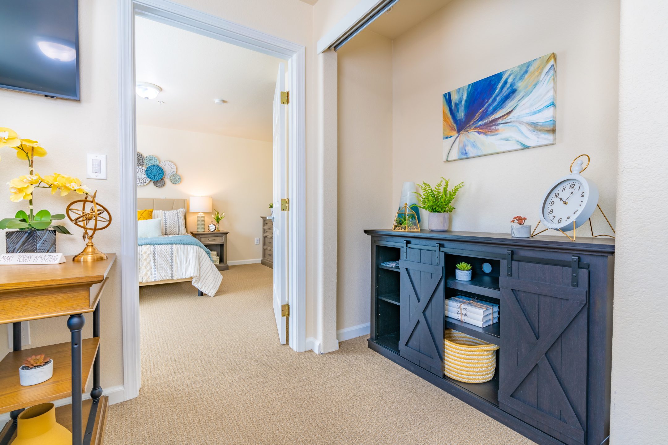 A bright hallway with beige carpet, modern decor, plants, and shelving creates an inviting path to a cozy Independent Living bedroom with a neatly made bed and yellow accents. Artwork and flowers add color to the space.