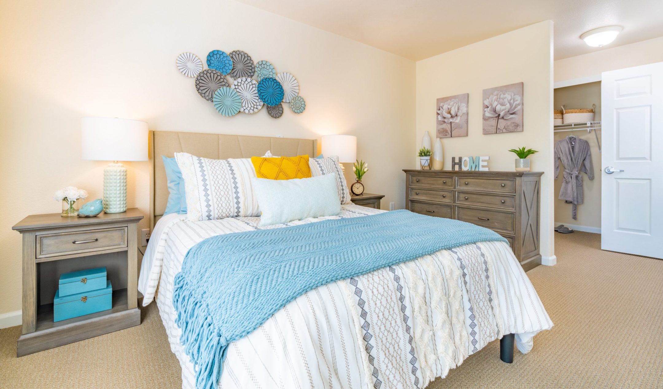 Bright bedroom with a neatly made bed in blue, white, and yellow tones, matching nightstands with lamps, decorative wall art, a dresser with plants, and an open closet door in the background.