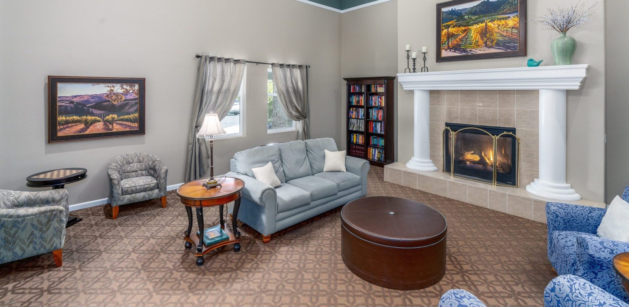 A cozy Independent Living living room with a light gray sofa, patterned chairs, a round coffee table, a fireplace with a fire, a tall bookshelf, and large landscape paintings on beige walls under a high ceiling with a chandelier.