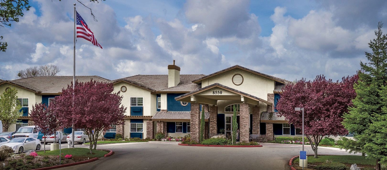 A large, two-story Eskaton Granite Bay senior living building with an entrance canopy, surrounded by blooming trees and parked cars, with an American flag flying in front under a partly cloudy sky.