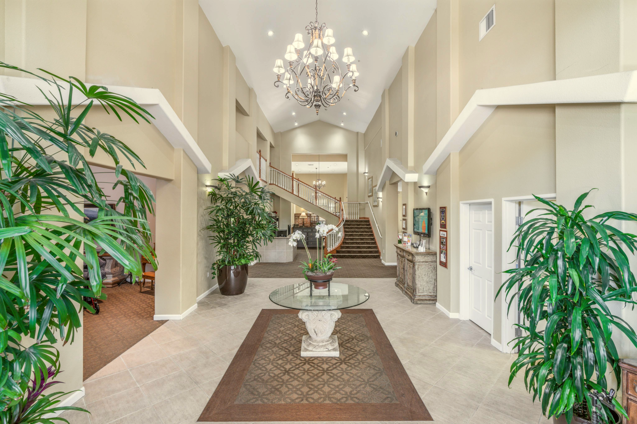 Spacious, elegant lobby with tall ceilings, a chandelier, large potted plants, and a round glass table with flowers welcomes you to your Eskaton Granite Bay tour; neutral walls and a carpeted staircase lead to an inviting upper level.