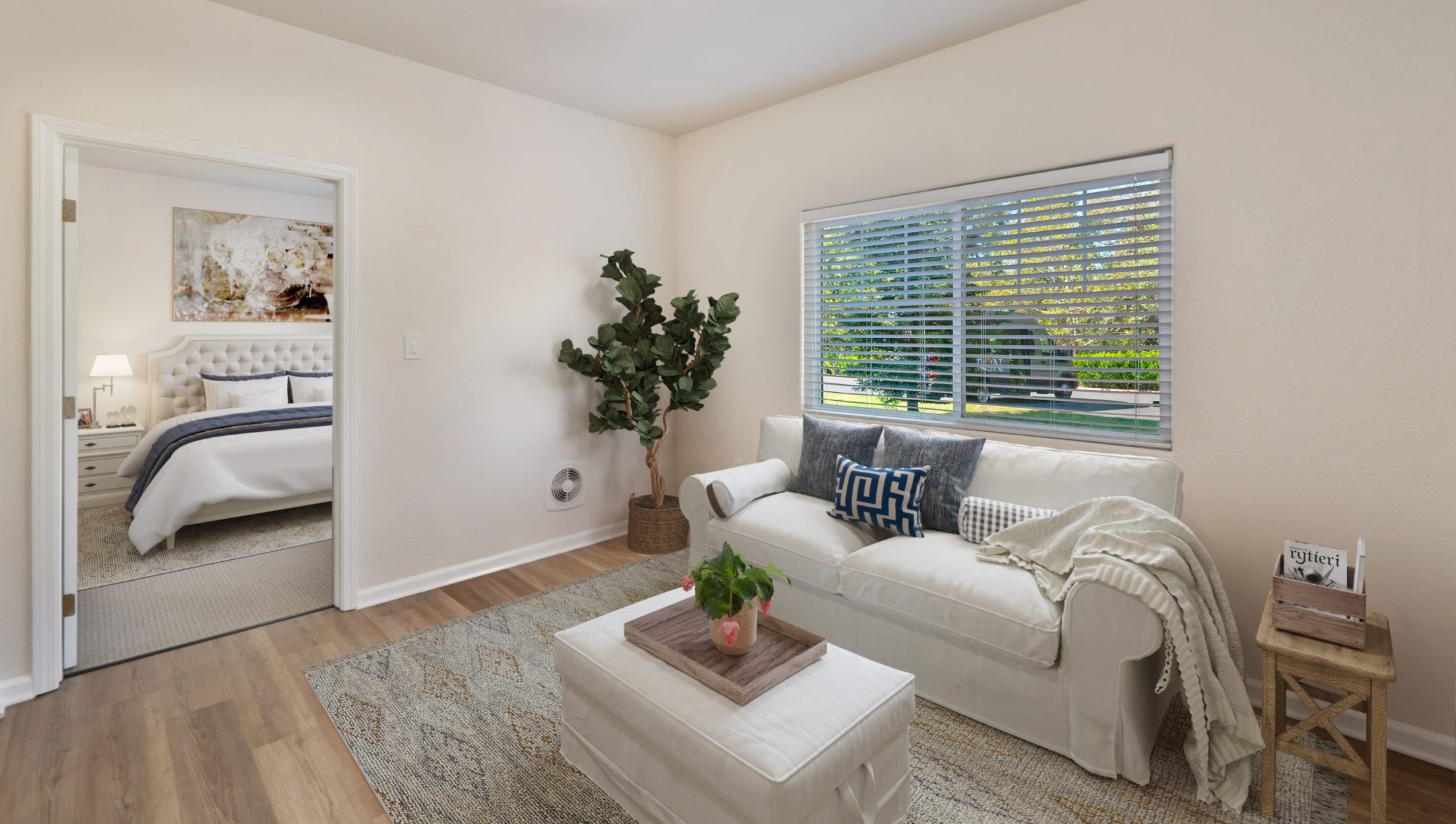 A cozy living room at Eskaton Granite Bay, perfect for short-term stays, with a white sofa, decorative pillows, a potted plant, and a small table with books. Natural light streams through window blinds, and a bedroom is visible through an open door.