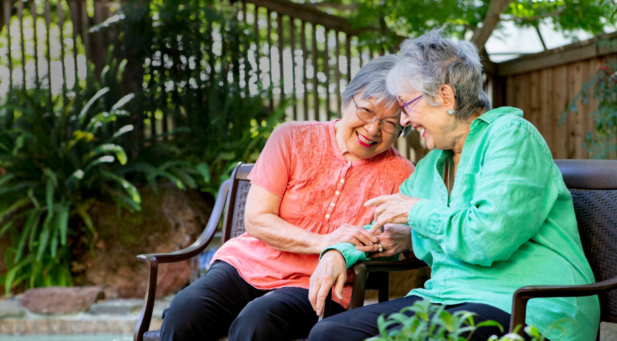 Two elderly women sit on a bench in a garden by a pool, smiling and laughing together. One wears a coral blouse and glasses, the other wears a teal blouse. They appear joyful and are enjoying each others company outdoors.