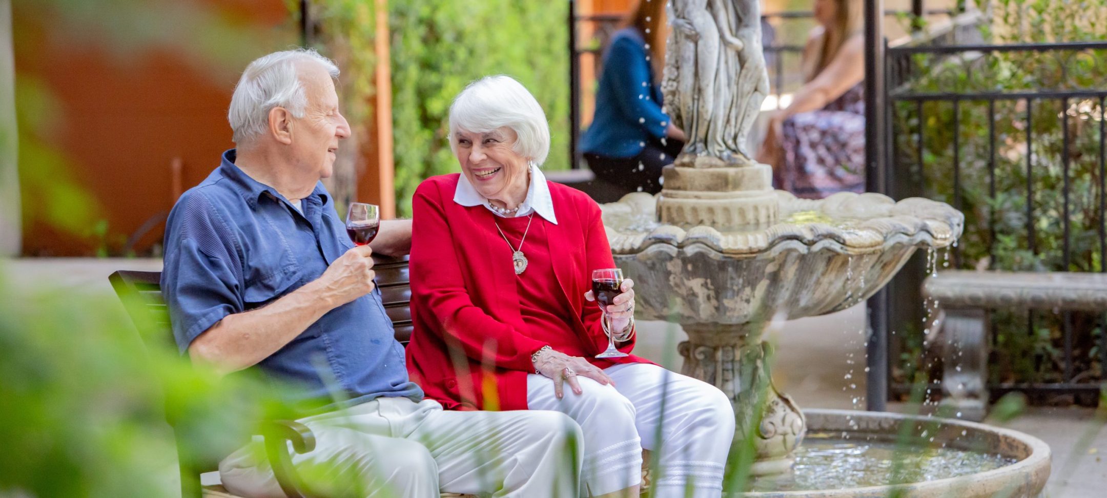 An elderly man and woman sit on a bench by a fountain, smiling and holding glasses of red wine, enjoying a pleasant outdoor conversation in a garden setting.