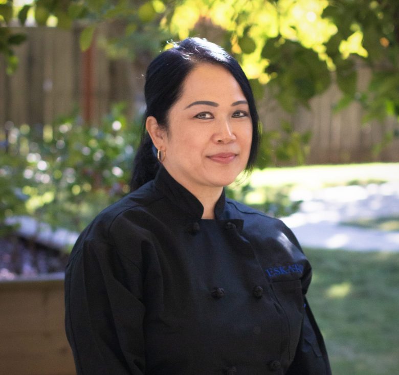 A woman with long dark hair wearing a black chef’s coat stands outdoors in a sunlit garden, smiling gently at the camera. Green foliage and a wooden fence are visible in the blurred background, reflecting the fresh vibe of Eskaton Land Park dining.