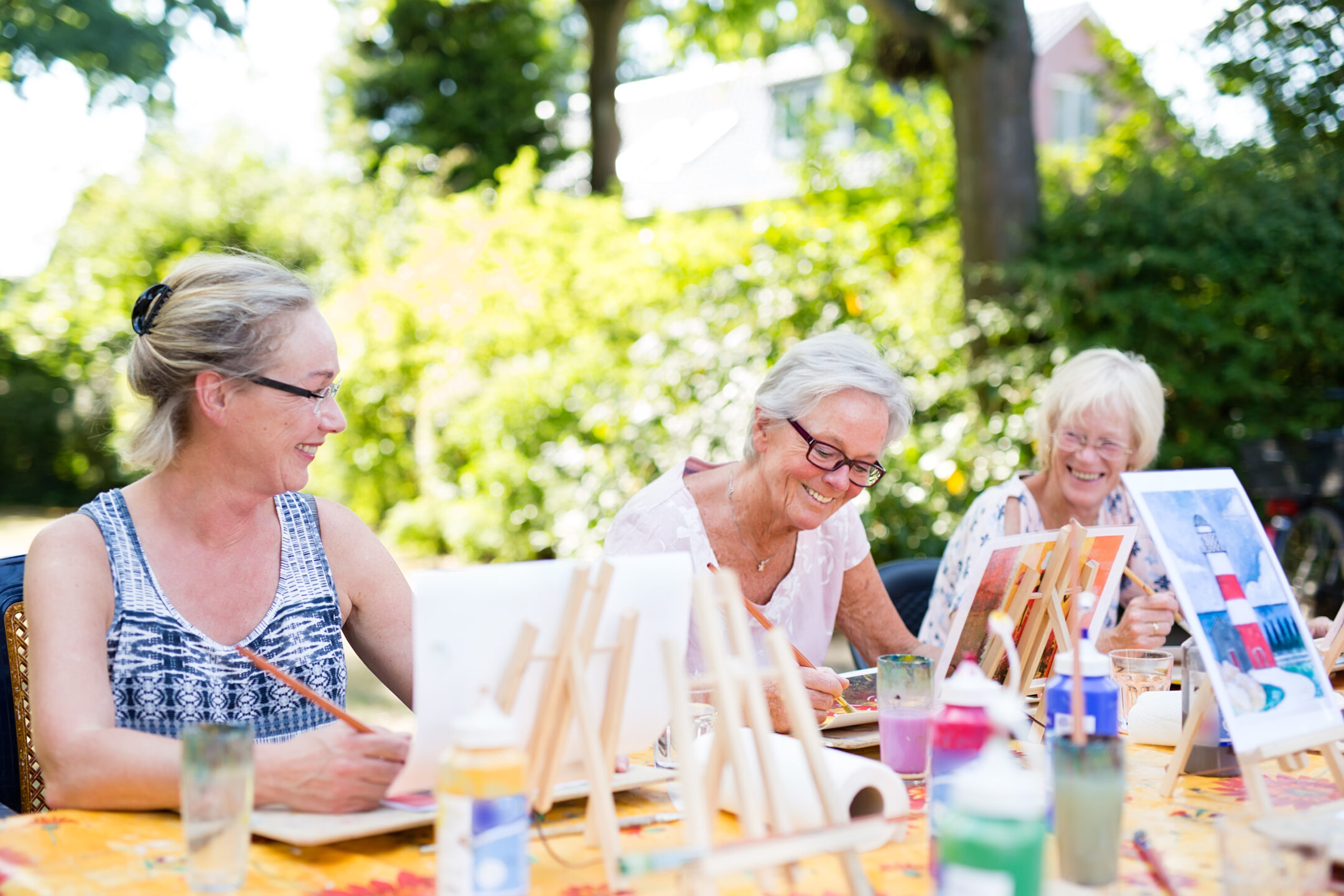 Three older women sit at a table outdoors, smiling and painting on canvases during a relaxing Petals & Pours afternoon paint event. Art supplies and drinks are scattered on the table, surrounded by trees and sunshine.