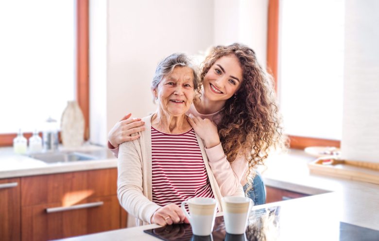 A smiling elderly woman and a young woman with curly hair embrace in a bright kitchen, standing behind a counter with two mugs. Sunlight streams in through large windows, creating a welcoming homepage feel.