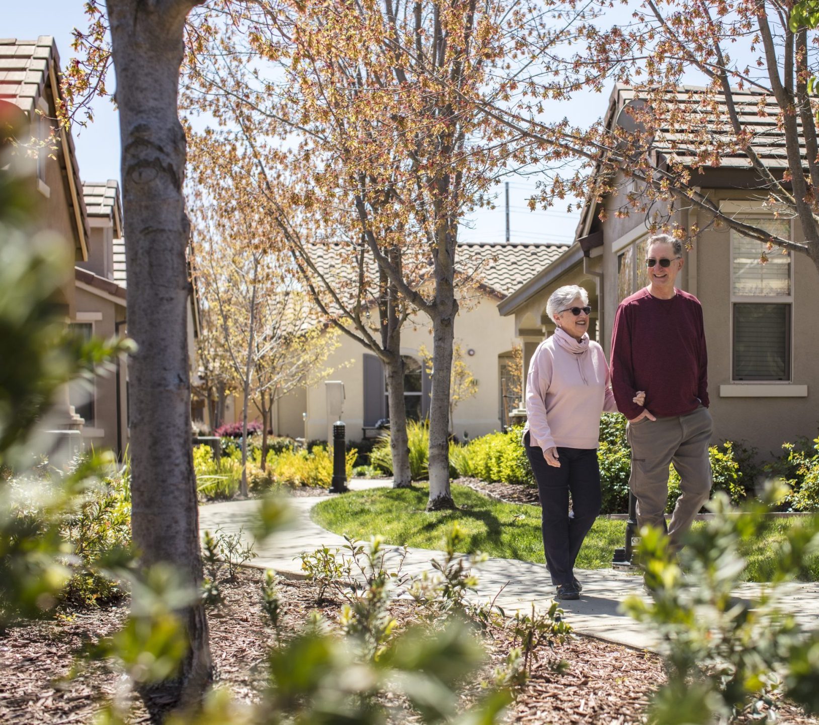An older woman and man walk together on a paved path through a landscaped neighborhood with small trees, green bushes, and single-story houses on a sunny day.