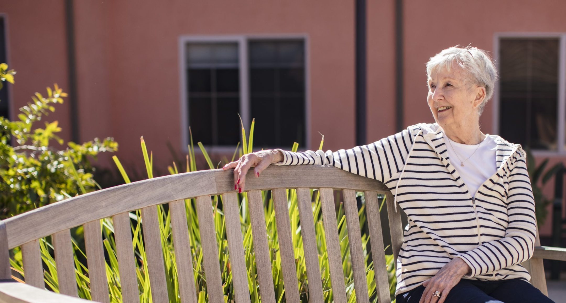 An older woman with short gray hair, wearing a striped hoodie and dark pants, sits smiling on a wooden bench outside, with green plants and a peach-colored building in the background.