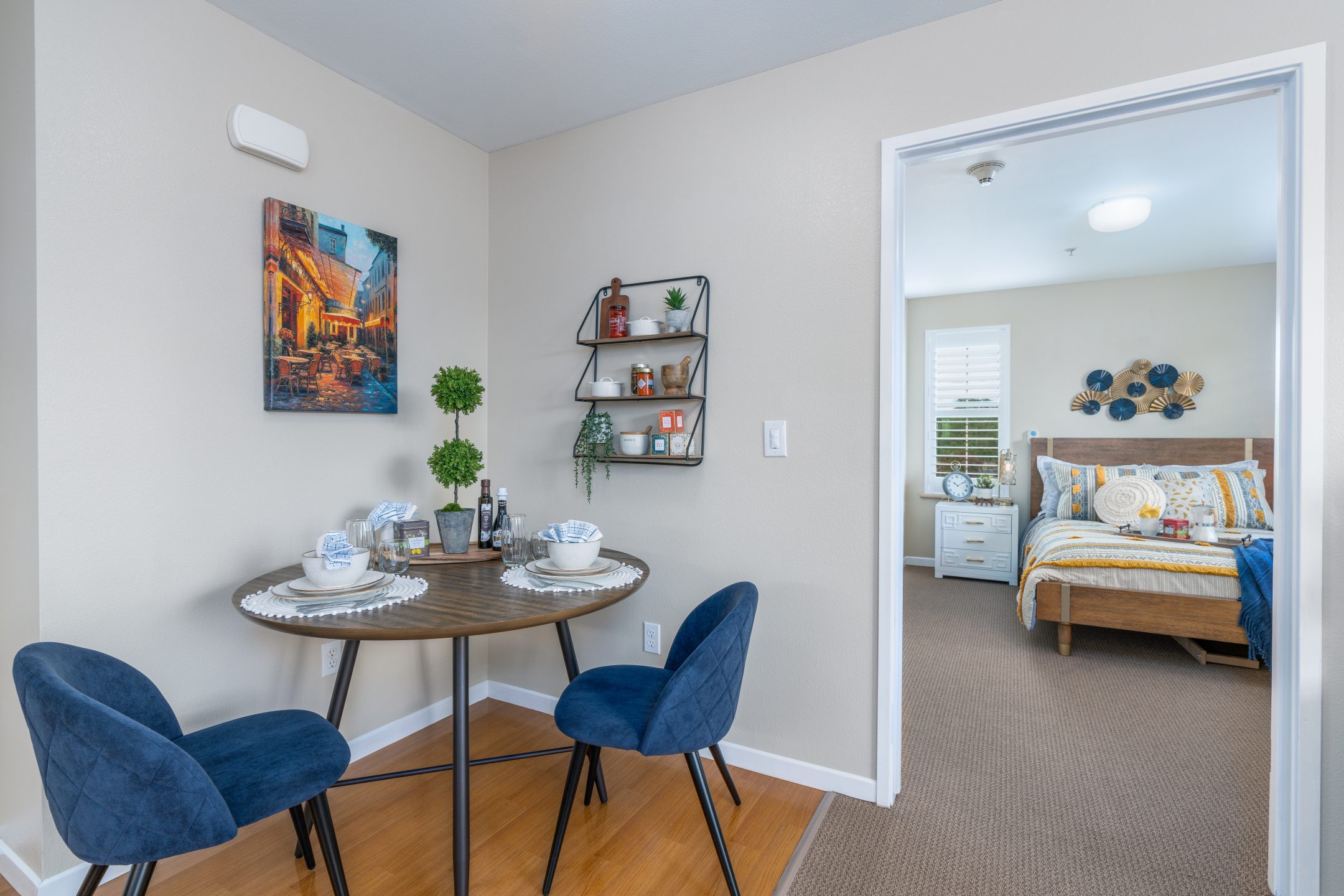 A small dining area with a round table set for two, blue velvet chairs, wall shelves with decor, and a view into a neatly made bedroom with natural light.