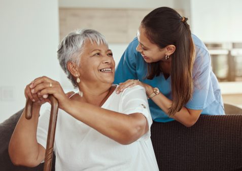 An elderly woman with gray hair sits on a couch, smiling and holding a cane, while a young female caregiver in scrubs from Eskaton Village Placerville senior living stands behind her, gently touching her shoulders and smiling warmly.
