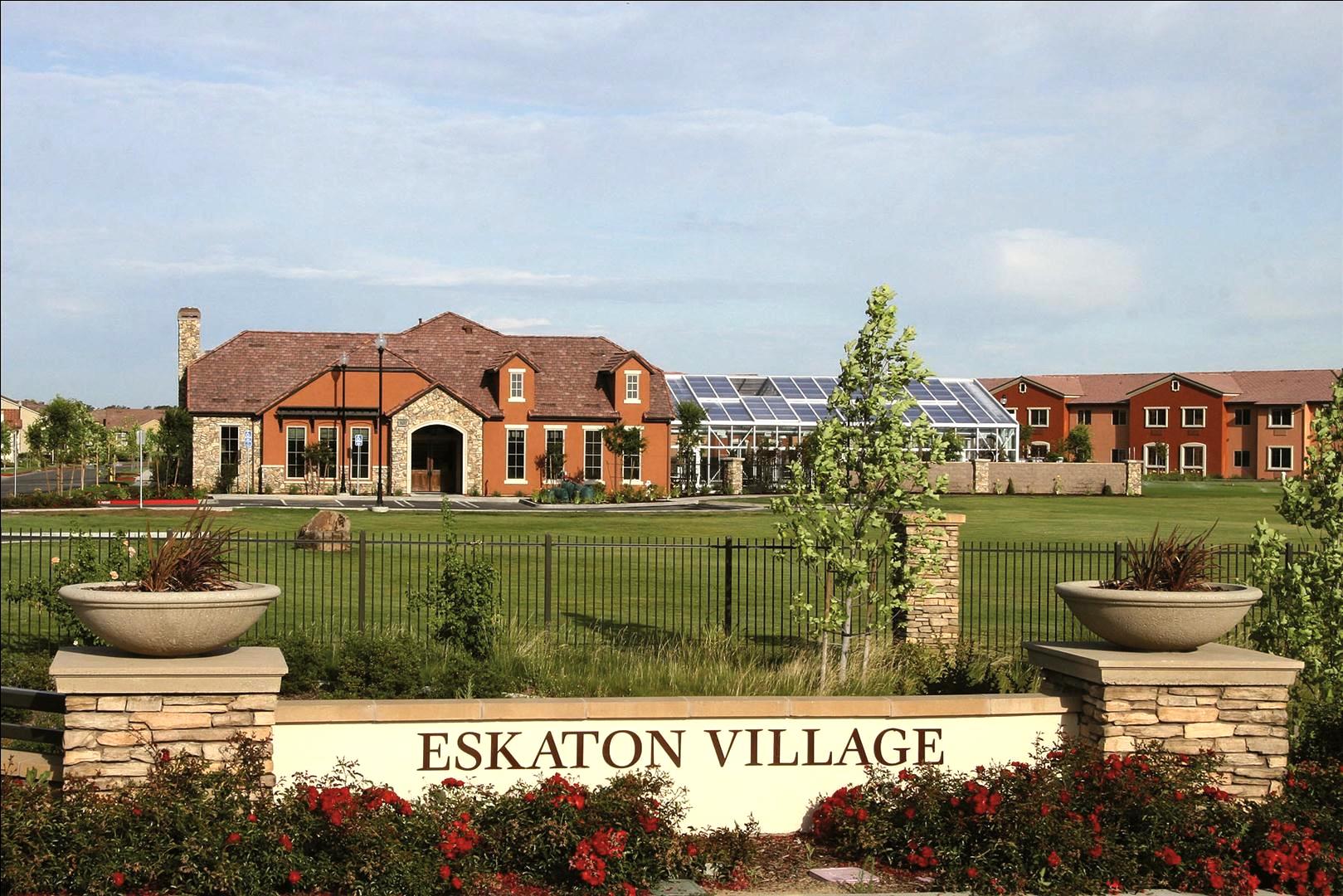 A large, modern building complex labeled Eskaton Village sits behind a landscaped area with red flowers and stone planters, surrounded by a black fence under a clear sky.