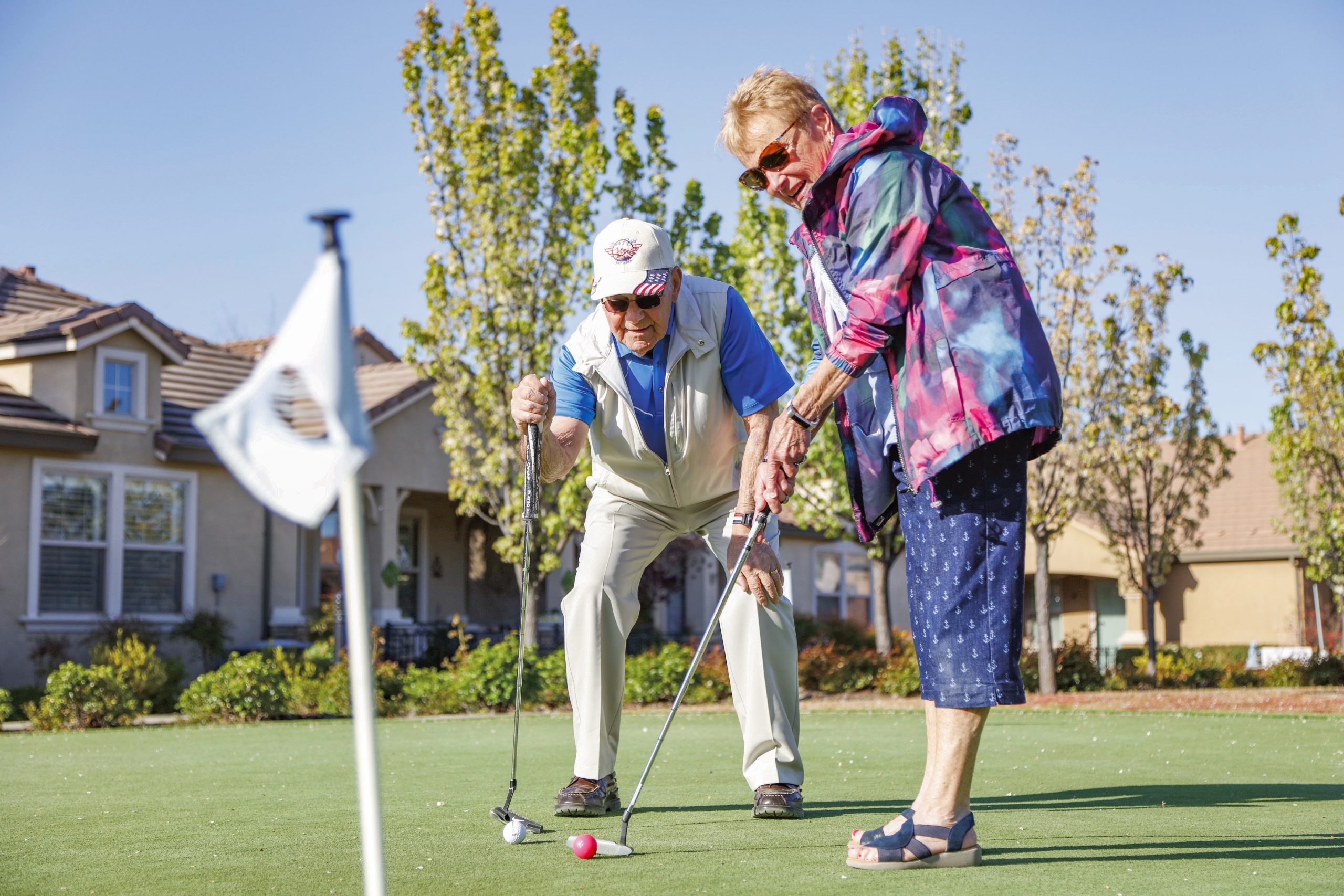 An older man and woman play mini golf outside on a sunny day. The man, wearing a cap and light vest, points to help the woman aim her putt. Houses and trees are in the background near the golf green.