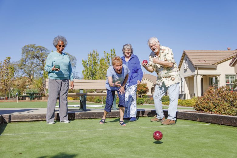 Four smiling seniors play bocce ball on a sunny day in a park, with houses and trees in the background. One woman is throwing a ball while the others watch and cheer.