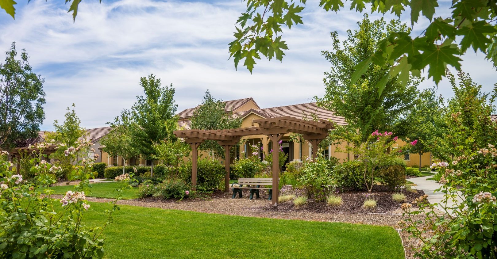 A landscaped garden with green grass, leafy trees, flowering bushes, and a wooden pergola over a bench, with yellow buildings and a partly cloudy sky in the background.