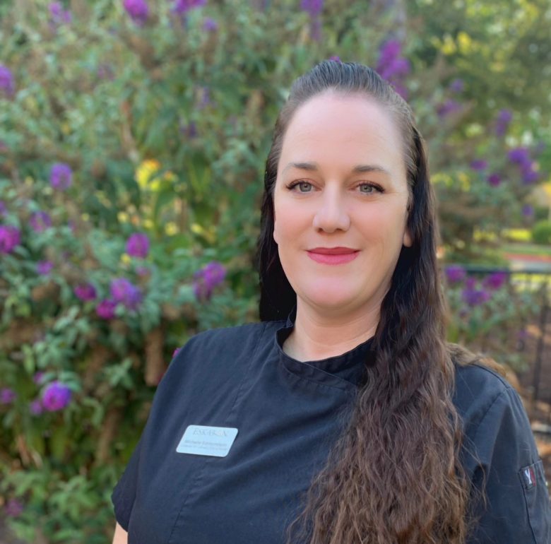 A woman with long brown hair, wearing a black shirt and name tag, stands outside in front of green bushes with purple flowers, smiling at the camera—representing Eskaton Village Placerville Culinary.