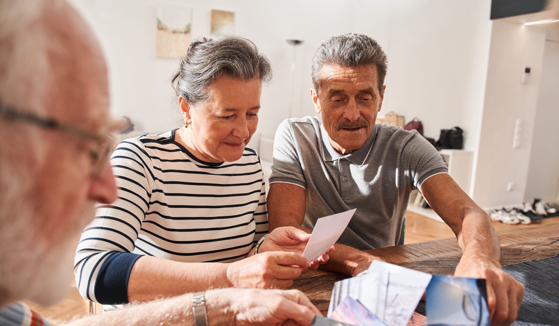Three older adults sit at a table indoors, smiling and looking at photographs together. Sunlight streams into the room, creating a warm and cozy atmosphere.