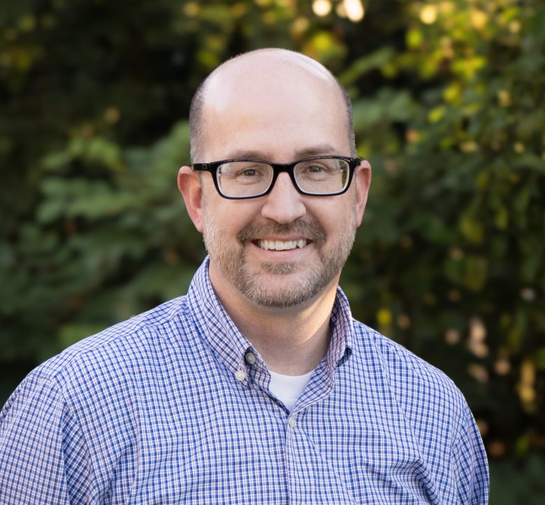 A smiling man with glasses, a beard, and a bald head, wearing a blue and white checkered shirt, stands outdoors in front of green foliage at Eskaton Village Roseville senior living.