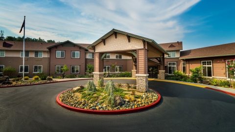 A modern assisted living facility with brown siding, covered entrance, landscaped circular driveway with rocks and small trees, and a flagpole on the left under a partly cloudy sky.
