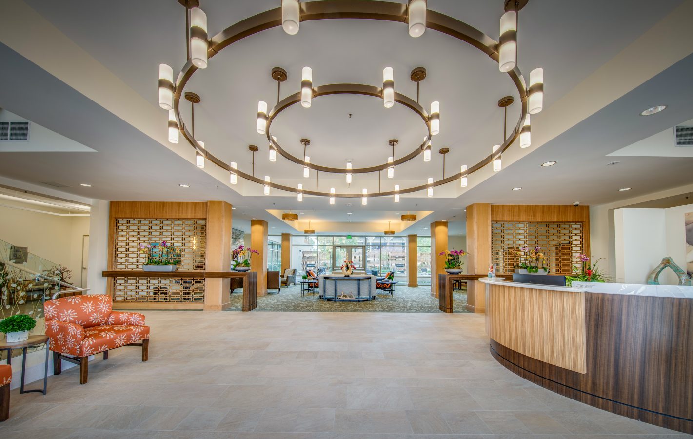 Spacious, modern hotel lobby with a large circular chandelier, contemporary seating area, reception desk, decorative plants, and large windows letting in natural light.