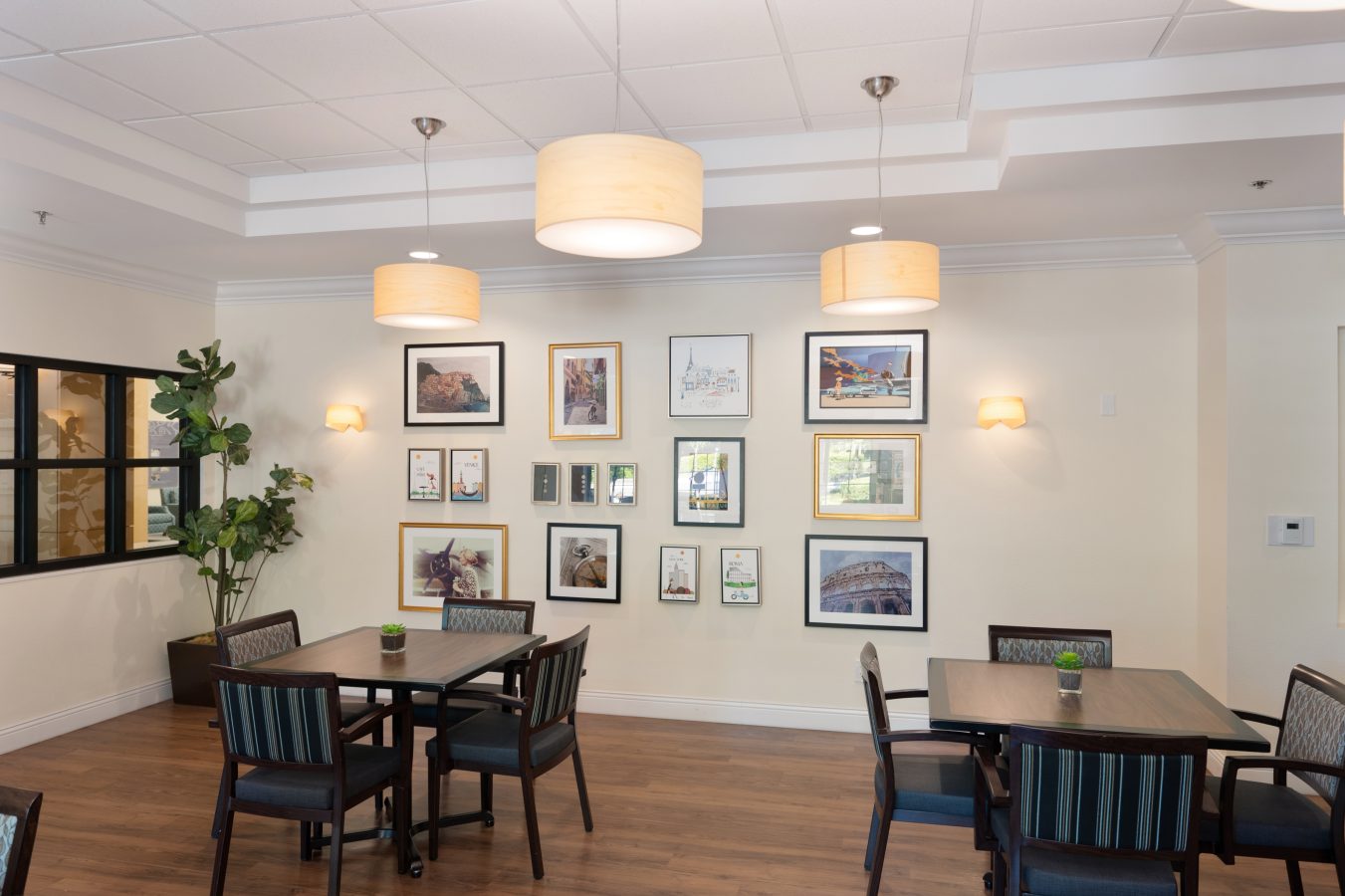 A bright dining area with wooden floors, modern pendant lights, framed artwork on the wall, potted plants, and tables with striped chairs arranged for group seating.