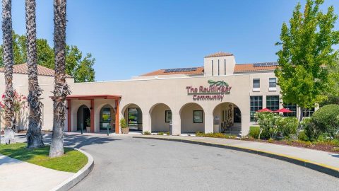 The exterior of a building labeled “The Redwoods Community,” featuring arched entryways, palm trees, and landscaping under a clear blue sky.