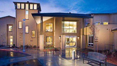 Modern building with large windows and warm lighting at dusk, featuring a tall entryway, landscaped garden, wet pavement, and a metal bench in the foreground.