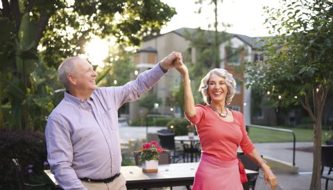 An older man and woman smile and dance together outdoors near a table with flowers, with trees and a building in the background on a sunny day.