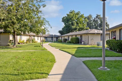 A curved sidewalk runs through a grassy courtyard surrounded by single-story apartment buildings, with trees, shrubs, and lamp posts lining the path under a partly cloudy sky.