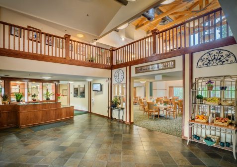 Spacious lobby with tiled floor, wood accents, a reception desk, wall clock, and open view of a dining area. A display shelves holds fresh fruit, and natural light brightens the inviting space.
